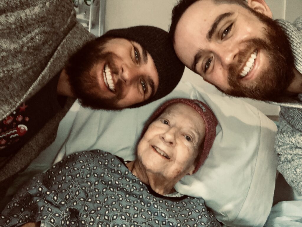 A grandmother in a hospital bed with her two (bearded) grandsons, wearing a winter hat and hospital gown, all three smiling :)