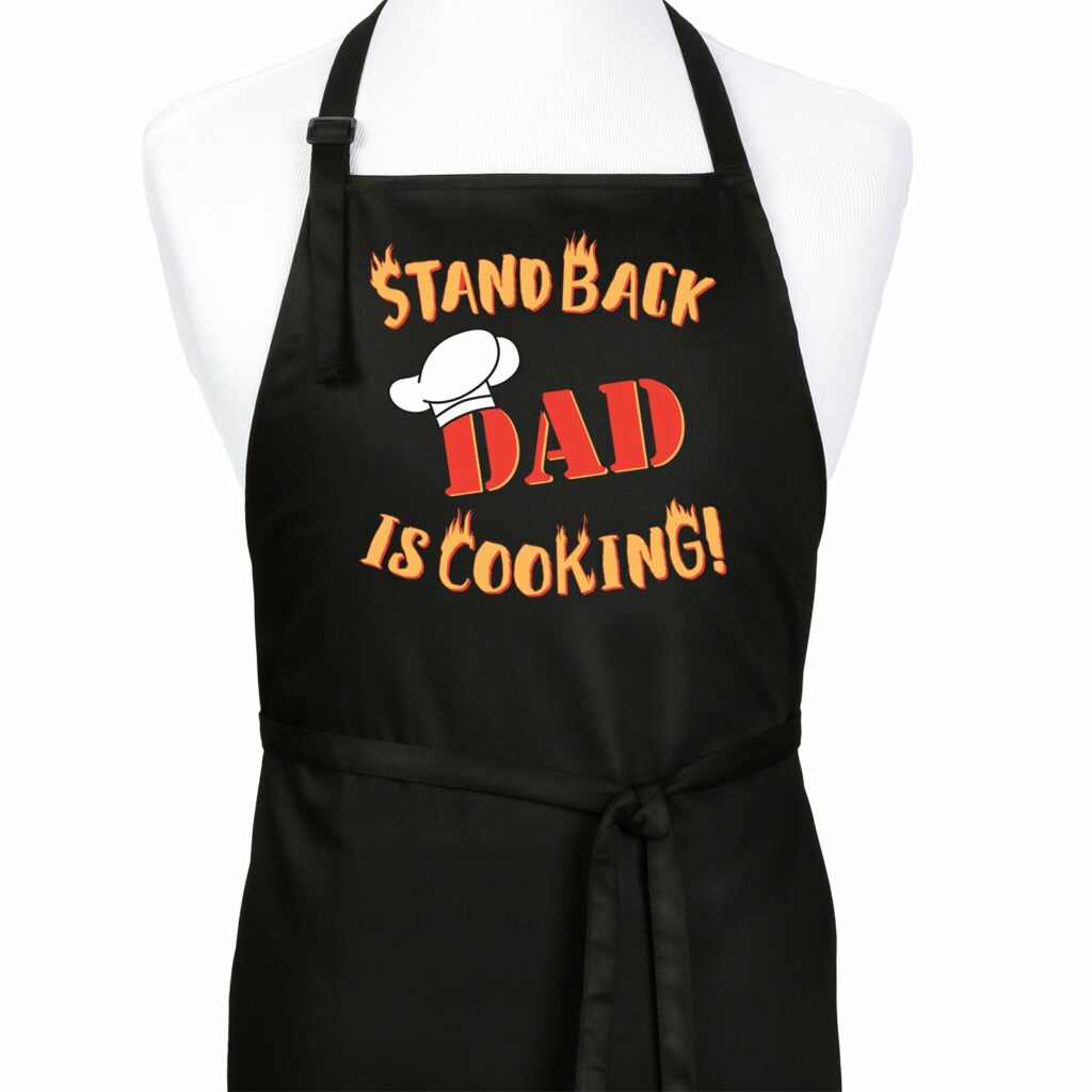 Black apron reading in yellow text "STAND BACK" and then in red text "DAD" with a chef hat on the first D in DAD, then below that "Is Cooking!" All of the yellow text appears to be on fire.